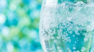 Obraz przedstawia zbliżenie na szklankę orzeźwiającej wody Culligan z licznymi pęcherzykami gazu, na błękitno-zielonym, rozmytym tle.