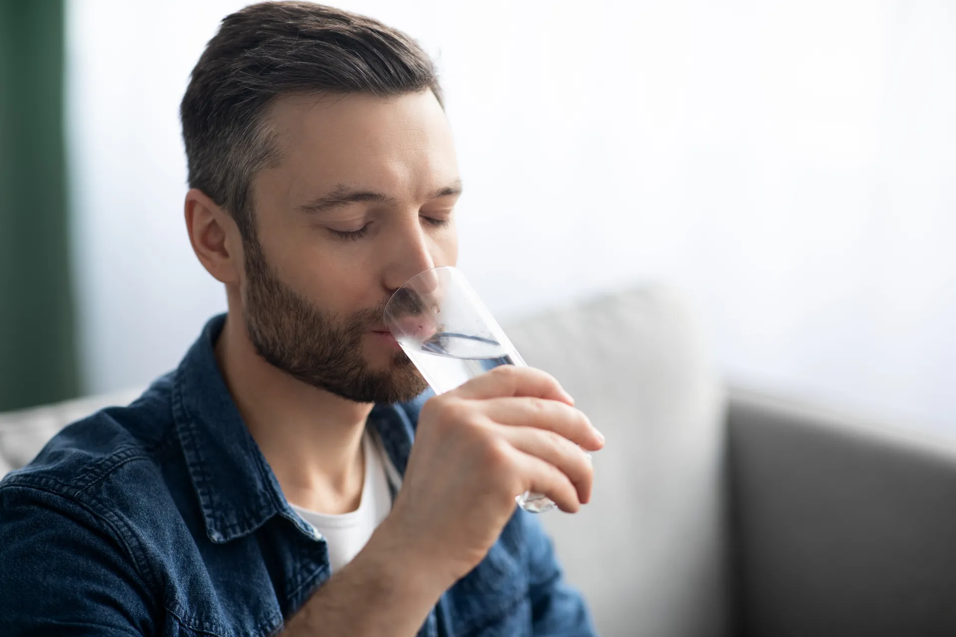 śmiechnięty, brodaty mężczyzna w dżinsowej koszuli pije orzeźwiającą szklankę czystej wody Culligan w jasnym pomieszczeniu.